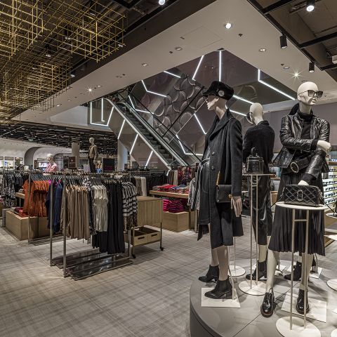 Ein modernes Interieur eines Bekleidungsgeschäfts mit Schaufensterpuppen in modischen Outfits. An den Ständern sind verschiedene Kleidungsstücke ausgestellt, im Hintergrund ist eine Rolltreppe zu sehen. Geometrisches Dekor und helle Beleuchtung werten den Raum auf.
