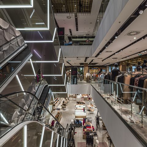 Innenansicht eines mehrstöckigen modernen Einkaufszentrums mit Rolltreppen und Glasgeländern. Auf den verschiedenen Etagen sind verschiedene Bekleidungsabteilungen zu sehen, die von Deckenleuchten beleuchtet werden. Moderne architektonische Details und Dekor schaffen eine elegante Atmosphäre.