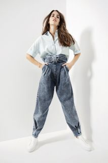 Eine Person posiert selbstbewusst in einer hellen, minimalistischen Umgebung. Sie trägt ein hellblaues Hemd mit Knopfleiste und hoch taillierte, plissierte Jeans mit weißen Turnschuhen. Die Jeans hat ein gerafftes Detail an der Taille.