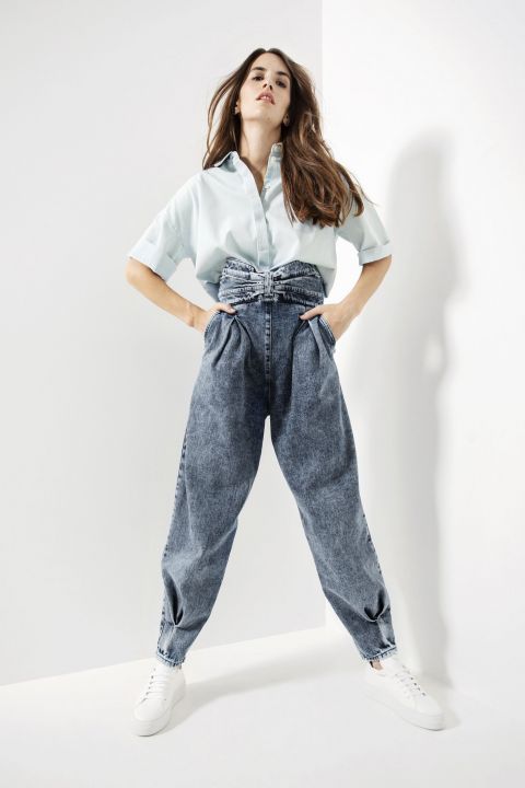 Eine Person posiert selbstbewusst in einer hellen, minimalistischen Umgebung. Sie trägt ein hellblaues Hemd mit Knopfleiste und hoch taillierte, plissierte Jeans mit weißen Turnschuhen. Die Jeans hat ein gerafftes Detail an der Taille.