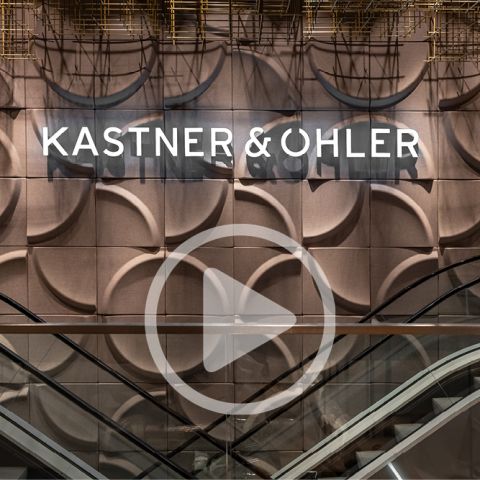 Ein moderner Innenraum zeichnet sich durch eine Wand mit kreisförmigen Mustern und dem beleuchteten Namen „KASTNER & ÖHLER“ aus. Darunter befinden sich zwei Rolltreppen, die in entgegengesetzte Richtungen führen. In der Mitte befindet sich eine transparente dekorative Auflage, die an eine Play-Taste erinnert.
