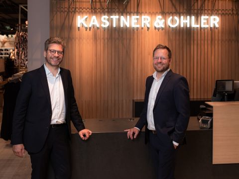 Zwei Männer in Anzügen stehen lächelnd an einem hölzernen Empfangstresen, an dessen Wand „KASTNER & ÖHLER“ leuchtet. Sie befinden sich in einem Innenbereich, wahrscheinlich in einem Geschäft oder Büro.