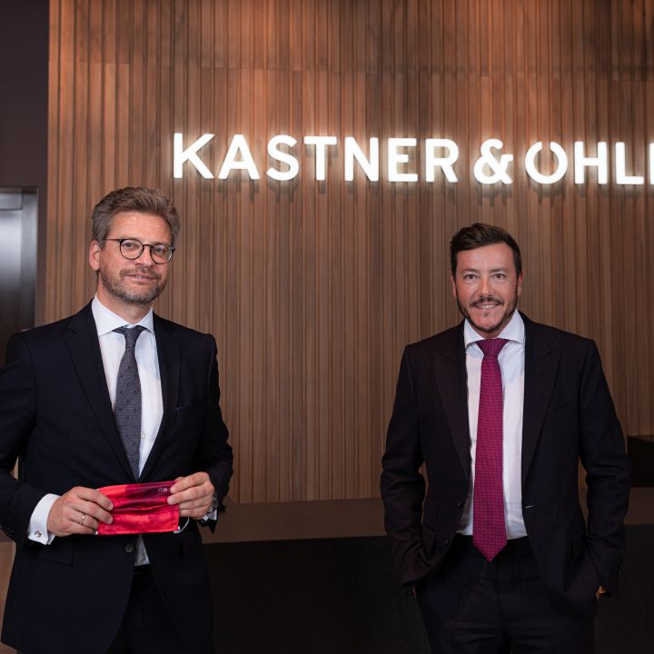 Zwei Männer in Anzügen stehen lächelnd vor einer Wand mit dem Leuchtschild „Kastner & Ohler“. Einer hält einen roten Mundschutz in der Hand. Im Hintergrund ist ein Holzpaneel-Design zu sehen.