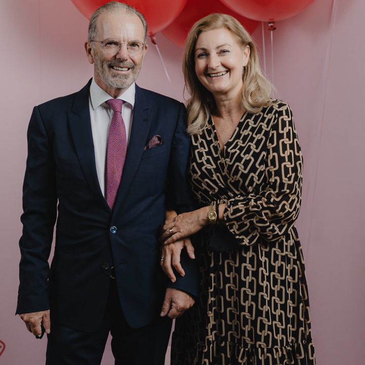 Ein lächelndes Paar steht vor großen roten Luftballons und einem rosa Hintergrund. Der Mann trägt einen dunklen Anzug und eine Krawatte, die Frau ein gemustertes Kleid. Sie halten Händchen und wirken glücklich und entspannt.
