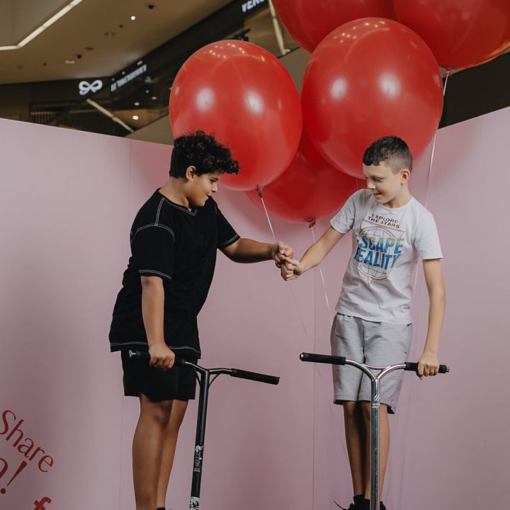 Zwei Jungen stehen auf Rollern, halten sich an den Händen und lächeln. Der Junge links trägt ein schwarzes Outfit, während der Junge rechts ein weißes Hemd und Shorts trägt. Sie halten große rote Luftballons und stehen vor einem rosa-weißen Hintergrund.