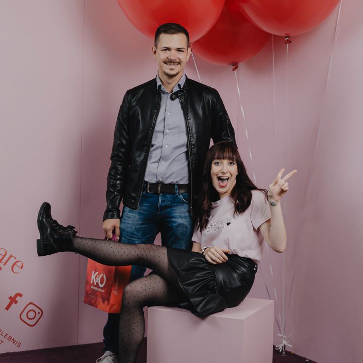 Ein Mann steht lächelnd vor einem Display mit roten Luftballons. Er trägt eine Lederjacke und Jeans. Eine Frau sitzt auf einem Würfel neben ihm, lächelt und macht ein Peace-Zeichen. Sie trägt ein rosa Hemd und einen schwarzen Rock mit gepunkteten Strumpfhosen.