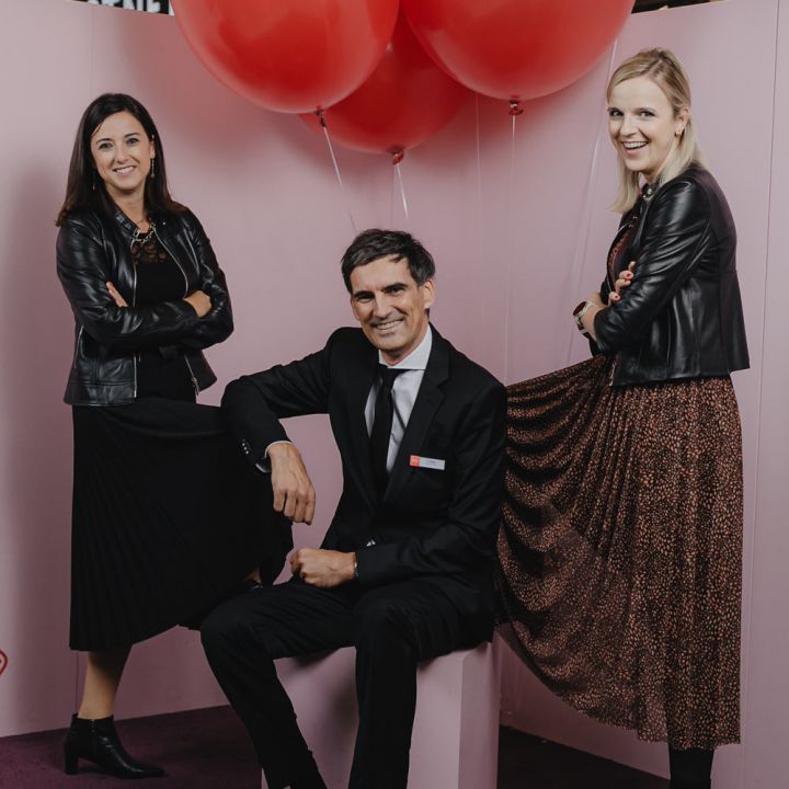 Drei Personen posieren vor einem rosa Hintergrund mit roten Luftballons. In der Mitte sitzt ein Mann im Anzug, flankiert von zwei Frauen in schwarzen Jacken. Sie lächeln und stehen auf einem lila Teppich.