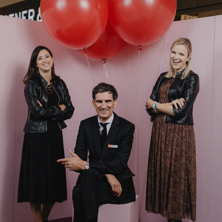 Drei Personen posieren vor einer rosa Wand mit großen roten Luftballons. Zwei Frauen stehen auf beiden Seiten eines sitzenden Mannes. Sie sind schwarz gekleidet und lächeln in die Kamera. Die Kulisse hat eine festliche und professionelle Atmosphäre.