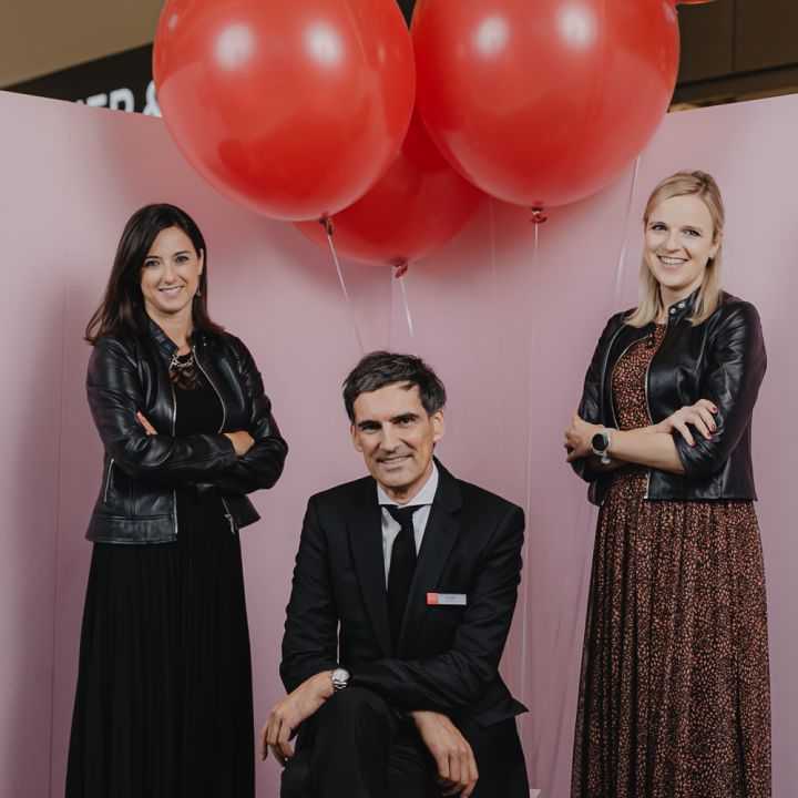 Drei Personen posieren vor einem rosa Hintergrund mit roten Luftballons. Zwei Frauen in Schwarz stehen mit verschränkten Armen zu beiden Seiten eines Mannes im schwarzen Anzug, der in der Mitte kniet. Sie lächeln alle.