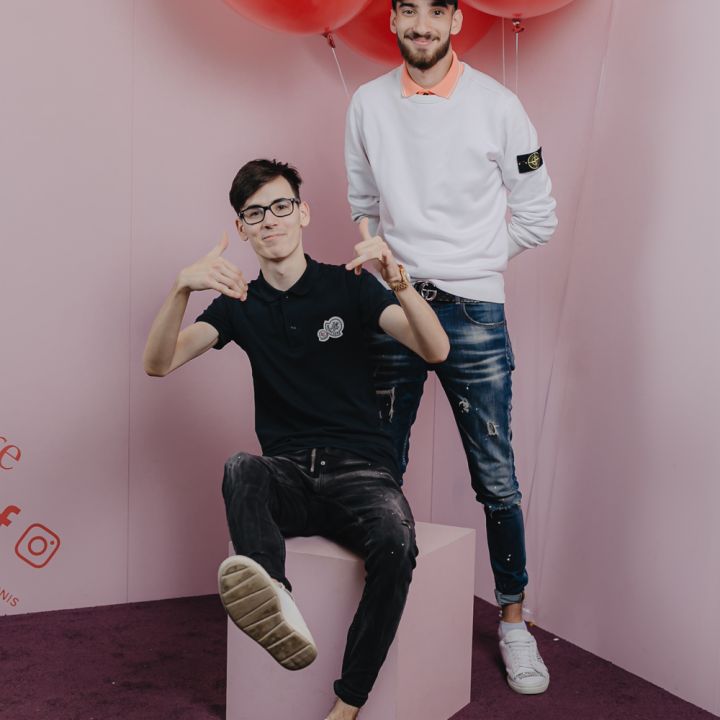 Zwei Männer posieren vor einem rosa Hintergrund mit großen roten Luftballons. Ein Mann im schwarzen Hemd sitzt auf einem Würfel und gestikuliert mit den Händen, während der andere im weißen Hemd und mit Mütze lächelnd hinter ihm steht.