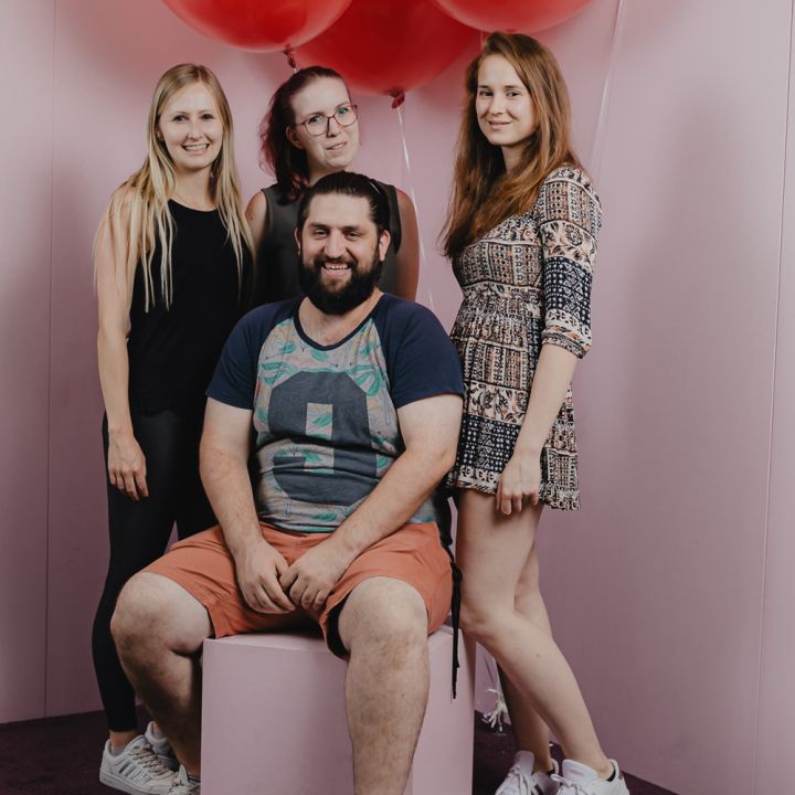 Eine Gruppe von vier Personen posiert vor einem rosa Hintergrund mit roten Luftballons. Drei Frauen stehen und ein Mann sitzt auf einem Würfel. Die Frauen lächeln und der Mann sitzt in der Mitte und trägt ein buntes T-Shirt und Shorts.
