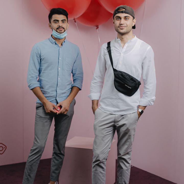 Zwei Männer stehen vor einem hellrosa Hintergrund mit drei großen roten Luftballons. Einer trägt ein hellblaues Hemd und graue Hosen, der andere ein weißes Hemd, graue Hosen und eine Mütze. Zwischen ihnen befindet sich eine kleine Plattform auf einem dunkelvioletten Teppich.