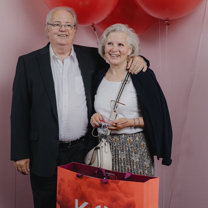 Älteres Paar lächelt und posiert vor großen roten Luftballons. Die Frau hält eine Einkaufstüte mit dem Aufdruck „K&Ö Kastner & Öhler“. Sie stehen vor einem rosa Hintergrund. Der Mann hat seinen Arm um die Schulter der Frau gelegt.
