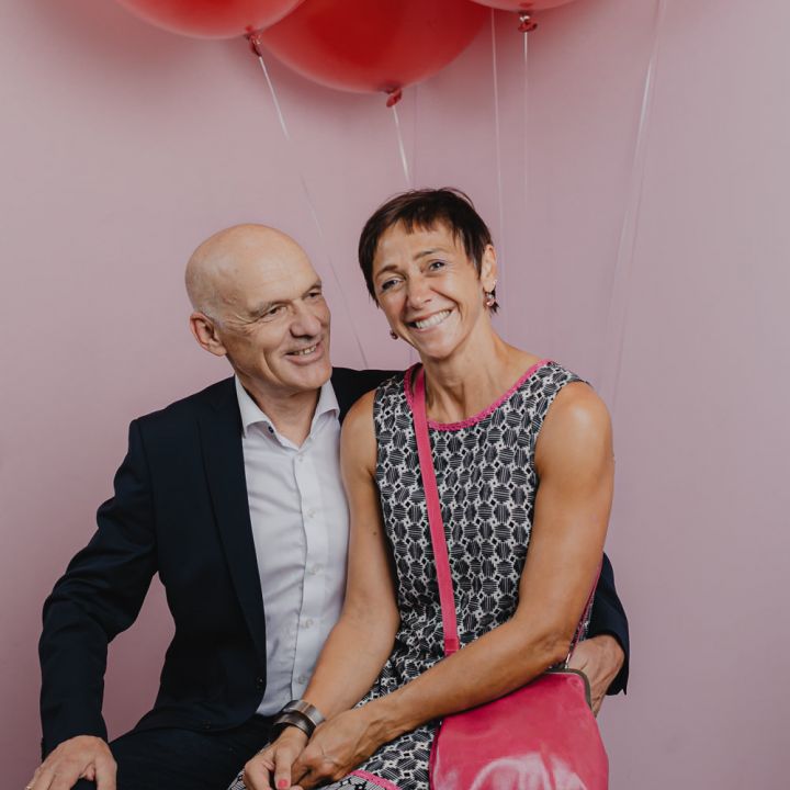 Ein lächelndes Paar posiert gemeinsam vor einem rosa Hintergrund mit roten Luftballons. Die Frau trägt ein gemustertes Kleid und eine rosa Handtasche, während der Mann im Anzug sitzt und seinen Arm um sie gelegt hat.