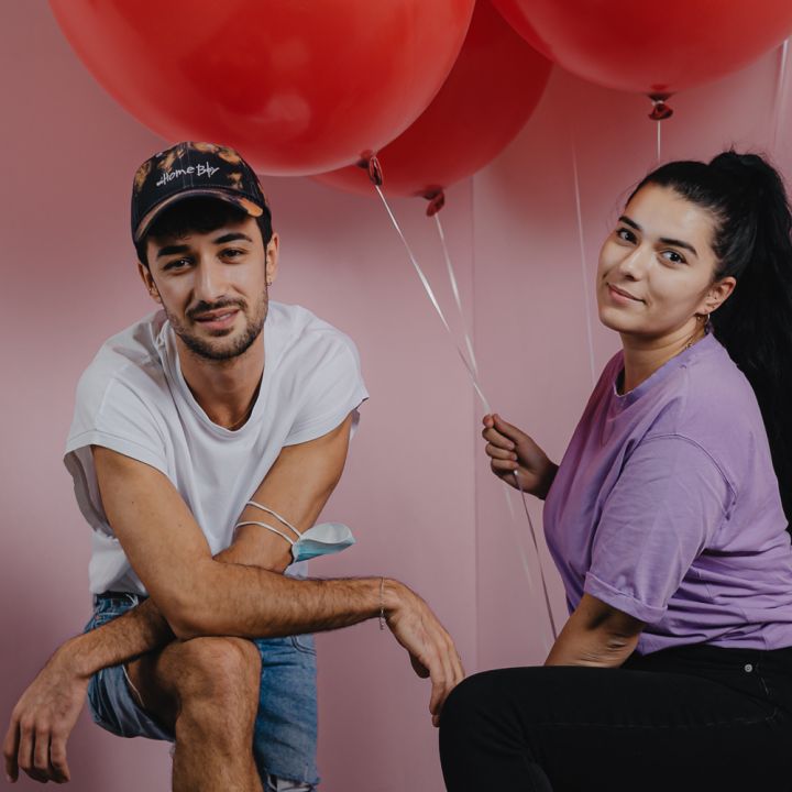 Ein Mann mit Mütze und eine Frau in lila Hemd posieren mit roten Luftballons vor einem rosa Hintergrund. Der Mann kniet leicht und lächelt, während die Frau sitzt und die Luftballonschnüre hält.