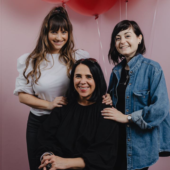 Drei Frauen posieren fröhlich vor rosa Wänden mit roten Luftballons über ihnen. Die Frau in der Mitte sitzt, während die anderen beiden lächelnd neben ihr stehen. Sie sind leger gekleidet und schaffen eine fröhliche und freundliche Atmosphäre.