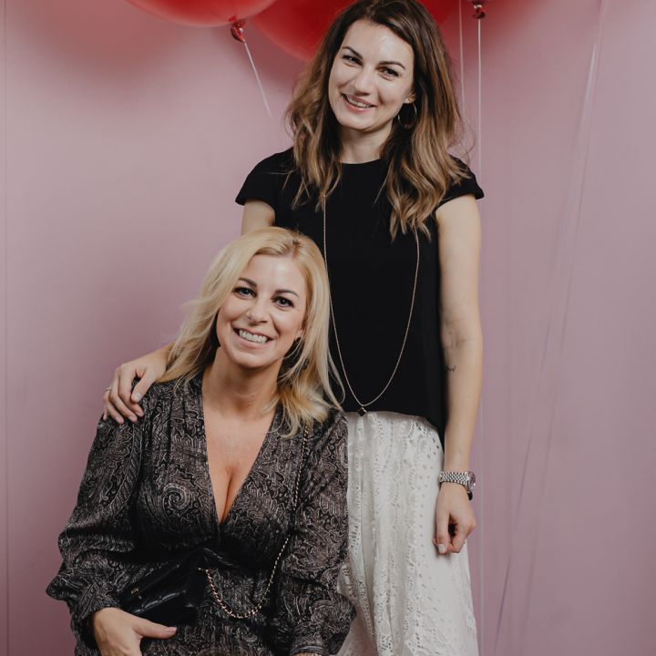 Zwei Frauen posieren vor rosa Wänden mit großen roten Luftballons darüber. Eine sitzt und trägt ein gemustertes Kleid, die andere steht in einem schwarzen Oberteil und weißen Hosen hinter ihr und lächelt, während sie ihre Hand auf der Schulter der sitzenden Frau legt.
