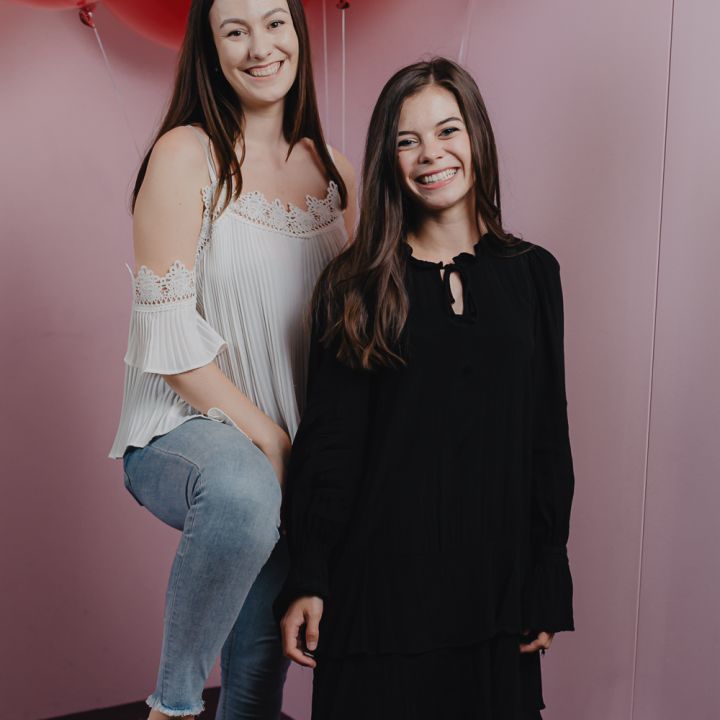 Zwei lächelnde Frauen vor einem rosa Hintergrund mit roten Luftballons. Eine sitzt und trägt ein weißes Oberteil und Jeans, während die andere steht und ein schwarzes Outfit trägt.