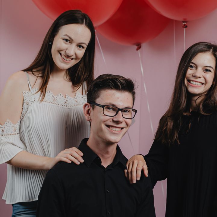 Drei Personen posieren vor roten Luftballons auf einem rosa Hintergrund. Ein Mann mit Brille sitzt da, hinter ihm stehen zwei Frauen. Sie lächeln alle und die Atmosphäre ist festlich und fröhlich.