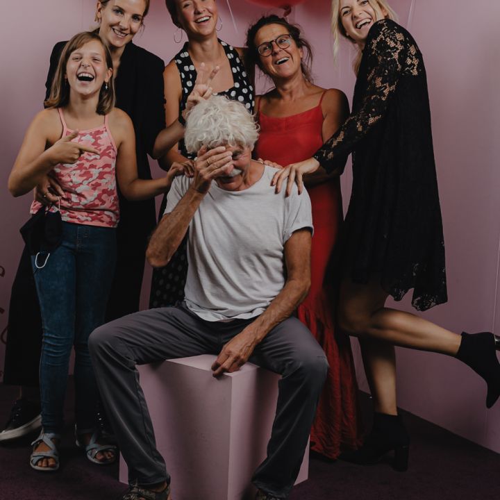 Eine Gruppe von sechs Personen posiert fröhlich vor einem rosa Hintergrund mit großen roten Luftballons. Eine Person sitzt auf einem rosa Würfel und verdeckt spielerisch ihr Gesicht, während die anderen lächelnd herumstehen und eine fröhliche Szene schaffen.