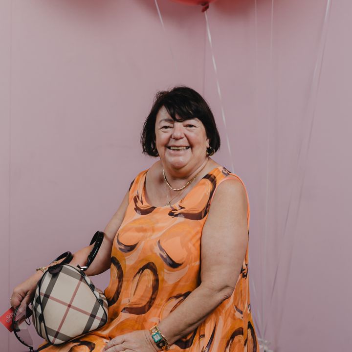 Eine Frau in einem orange gemusterten Kleid sitzt vor einer rosa Wand und hält eine karierte Handtasche in der Hand. Über ihr sind rote Luftballons zu sehen. Sie lächelt in die Kamera.