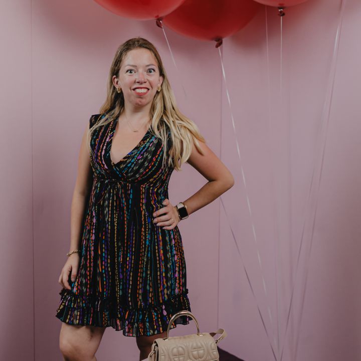 Eine Person in einem bunten Kleid steht selbstbewusst da, lächelt und hat die Hände in die Hüften gestemmt. Über ihr schweben große rote Luftballons vor einem rosa Hintergrund. Vor ihr steht auf einem Podest eine beige Handtasche.