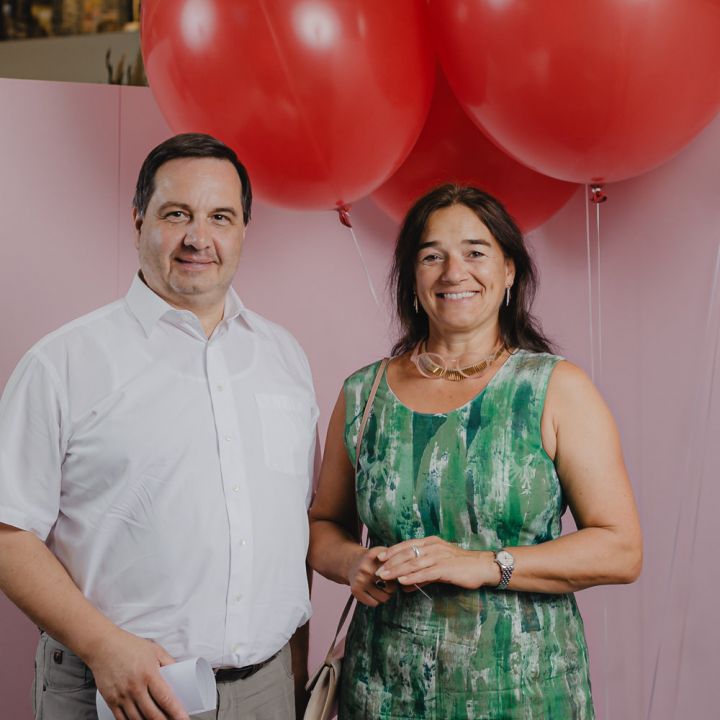 Ein Mann im weißen Hemd und eine Frau im grünen Kleid stehen lächelnd nebeneinander. Sie halten Papiere in den Händen und posieren vor einem hellrosa Hintergrund mit drei großen roten Luftballons.