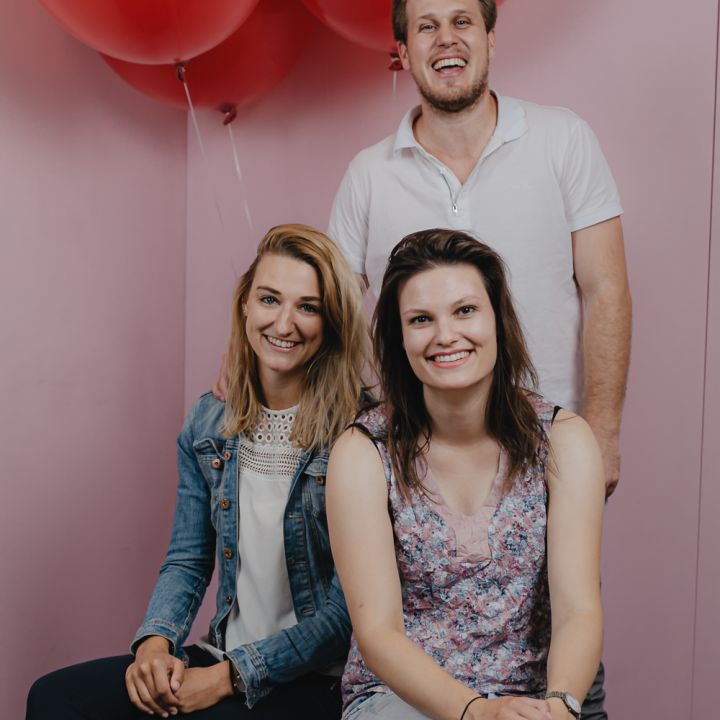 Drei Personen posieren in einem Studio mit roten Luftballons im Hintergrund. Zwei Frauen sitzen auf einer rosa Bank; die Frau links trägt eine Jeansjacke und die Frau rechts ein geblümtes Oberteil. Hinter ihnen steht ein lächelnder Mann in einem weißen Hemd.