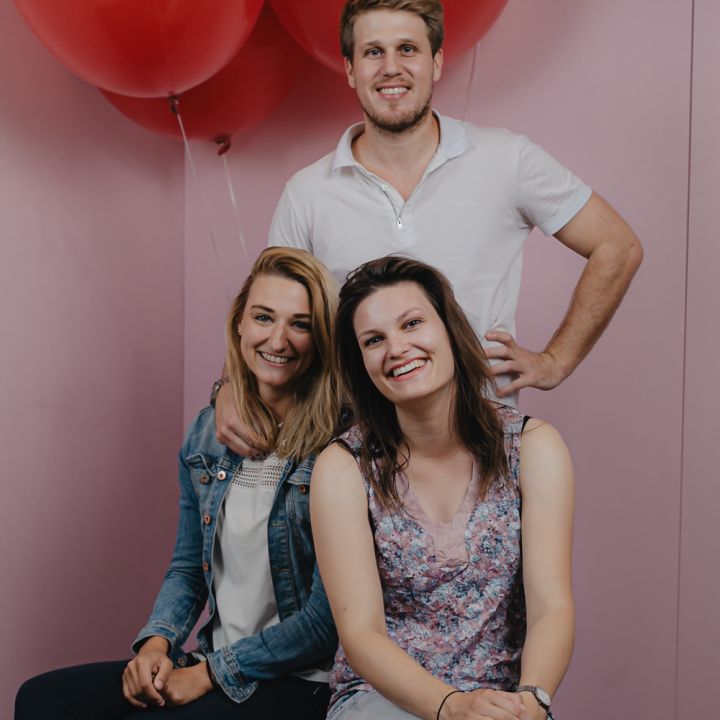 Drei Personen lächeln und posieren vor roten Luftballons vor einem rosa Hintergrund. Eine Person steht hinter den beiden anderen, die sitzen. Sie sind leger gekleidet und tragen einen Mix aus Jeans und heller Kleidung.