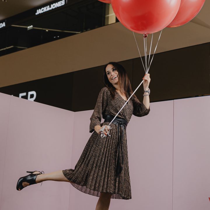 Eine Frau in einem schwarz-goldenen Kleid steht auf einer Plattform im Innenbereich und hält einen Haufen großer roter Luftballons. Sie lächelt und macht eine verspielte Pose mit einem angehobenen Bein. Sie trägt schwarze High Heels.