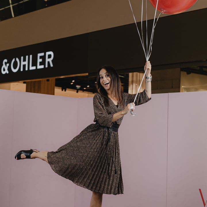 Eine Frau in einem gemusterten Kleid balanciert spielerisch auf einem Bein auf einer kleinen Plattform und hält dabei einen Haufen roter Luftballons in der Hand. Sie scheint sich in einem Einkaufszentrum zu befinden, im Hintergrund sind Ladenschilder zu sehen.