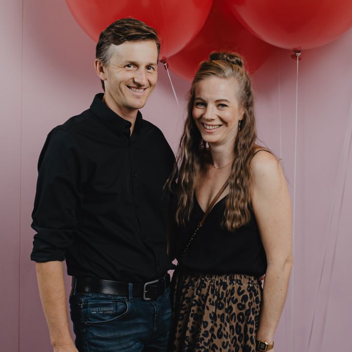 Ein Mann und eine Frau stehen lächelnd vor einem Hintergrund mit großen roten Luftballons. Der Mann trägt ein schwarzes Hemd und Jeans, die Frau ein schwarzes Top und einen Rock mit Leopardenmuster.