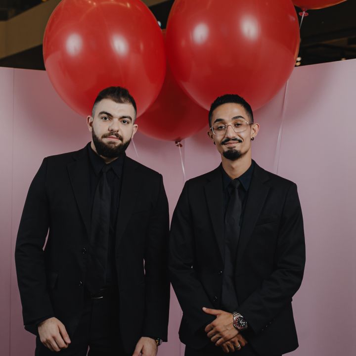 Zwei Männer in schwarzen Anzügen stehen vor einem rosa Hintergrund, die Hände hängen herab. Über ihnen schweben große rote Luftballons, die der Szene leuchtende Farben verleihen.