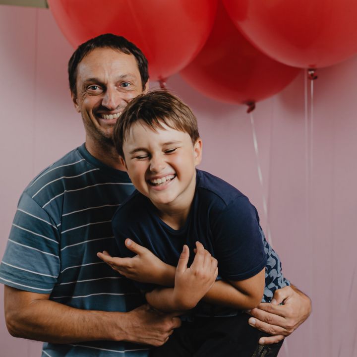 Ein Vater und sein Sohn lächeln fröhlich, wobei der Sohn spielerisch in den Armen des Vaters gehalten wird. Sie stehen vor mehreren großen roten Luftballons vor einem rosa Hintergrund. Der Sohn lacht mit geschlossenen Augen.