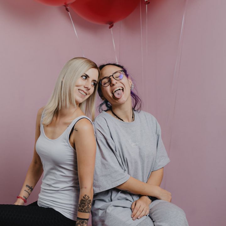 Zwei Personen sitzen und lächeln vor einem rosa Hintergrund mit roten Luftballons über ihnen. Eine Person hat blonde Haare und trägt ein weißes Tanktop; die andere hat lila Haare, eine Brille und trägt ein graues Outfit und streckt verspielt die Zunge heraus.