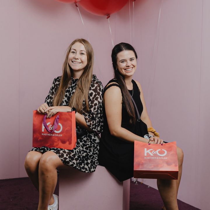 Zwei Frauen sitzen auf einer rosa Bank und halten jeweils eine rote Einkaufstüte in der Hand. Sie lächeln in die Kamera, umgeben von roten Luftballons. Im Hintergrund sind eine rosa Wand und ein lila Teppich zu sehen. Beide tragen lässige, stylische Outfits.