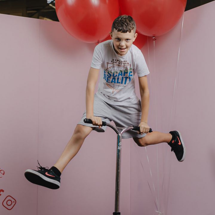 Ein Junge fährt fröhlich auf einem Roller durch die Luft vor einem pastellrosa Hintergrund. Er trägt ein hellgraues T-Shirt und Shorts sowie rote und schwarze Turnschuhe. Große rote Luftballons schweben hinter ihm und sorgen für eine verspielte Atmosphäre.