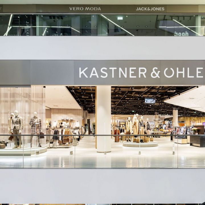 Eine moderne Ladenauslage bei Kastner & Öhler, in der Schaufensterpuppen in modischer Kleidung zu sehen sind. Das Geschäft verfügt über große Fenster, die einen gut beleuchteten Innenraum mit Kleiderständern und Accessoires in einem mehrstöckigen Einkaufszentrum freigeben.