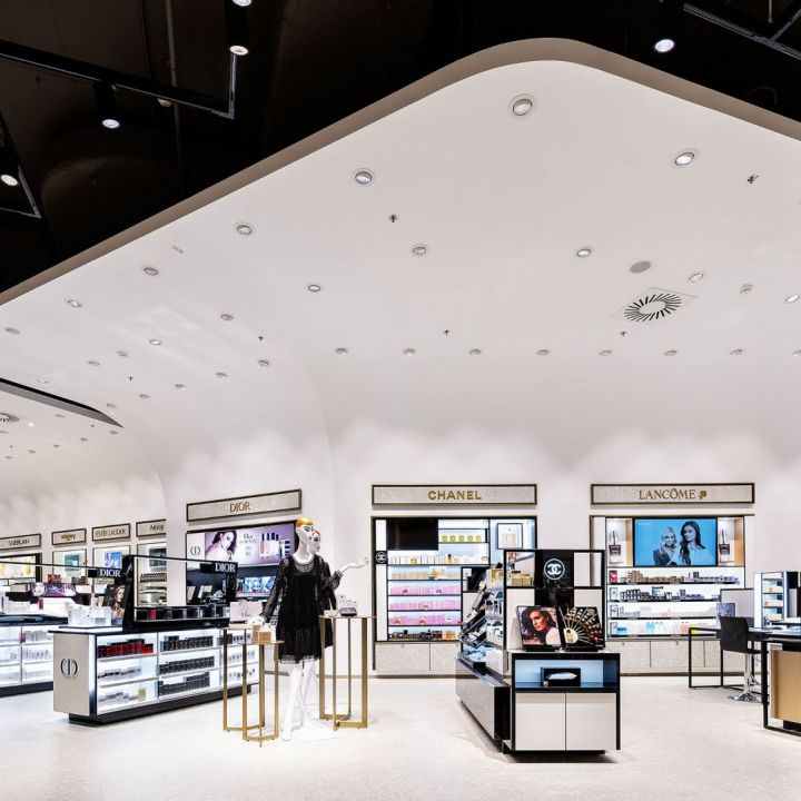 Ein moderner, hell erleuchteter Laden für Make-up und Kosmetik, in dem verschiedene Marken auf eleganten Regalen präsentiert werden. An der Decke befinden sich Strahler, und in der Mitte stehen Schaufensterpuppen in schwarzen Outfits.