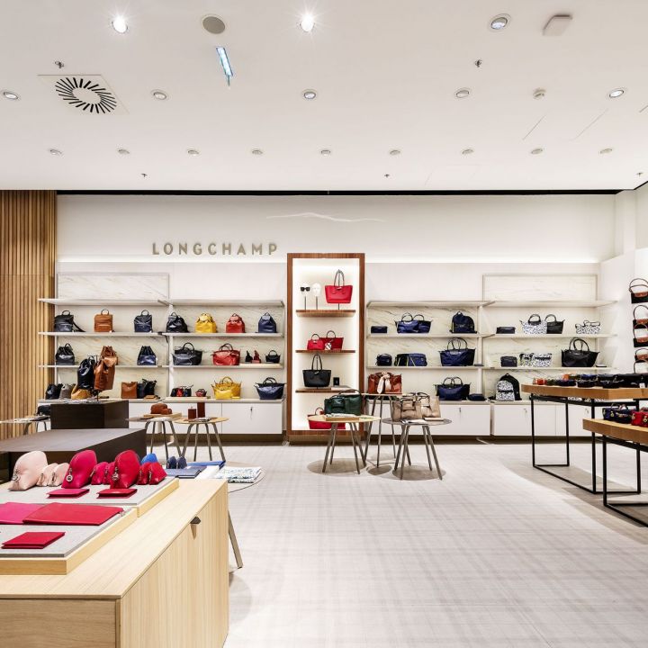 Ein modernes Interieur eines Longchamp-Geschäfts, in dem auf Regalen und Wänden eine Vielzahl von Handtaschen ausgestellt ist. Der Raum verfügt über einen hellen Boden, Tische mit Accessoires und eine helle Beleuchtung, die eine einladende Einkaufsatmosphäre schafft.