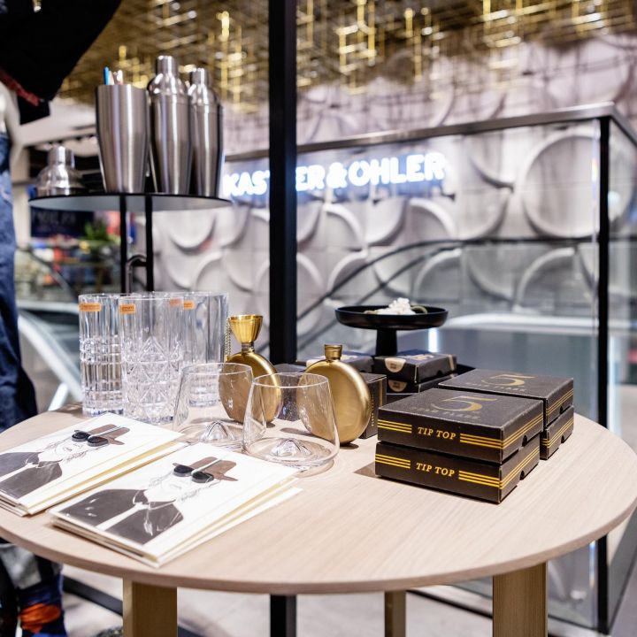 Auf einem Ausstellungstisch in einem Geschäft stehen Cocktail-Shaker, Whiskygläser, Bücher und ein Flachmann-Set. Im Hintergrund sieht man Kleidung, und das Ladendesign ist modern und stilvoll.