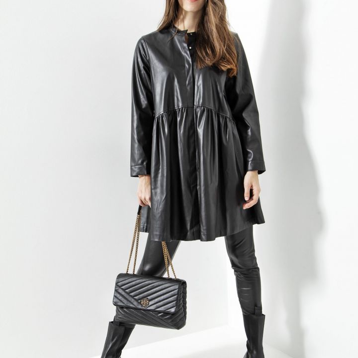 Eine Frau mit langen braunen Haaren steht vor einem weißen Hintergrund. Sie trägt ein schwarzes Lederkleid und Leggings. Sie hält eine schwarze gesteppte Handtasche und trägt schwarze Stiefeletten. Sie blickt selbstbewusst in die Kamera.