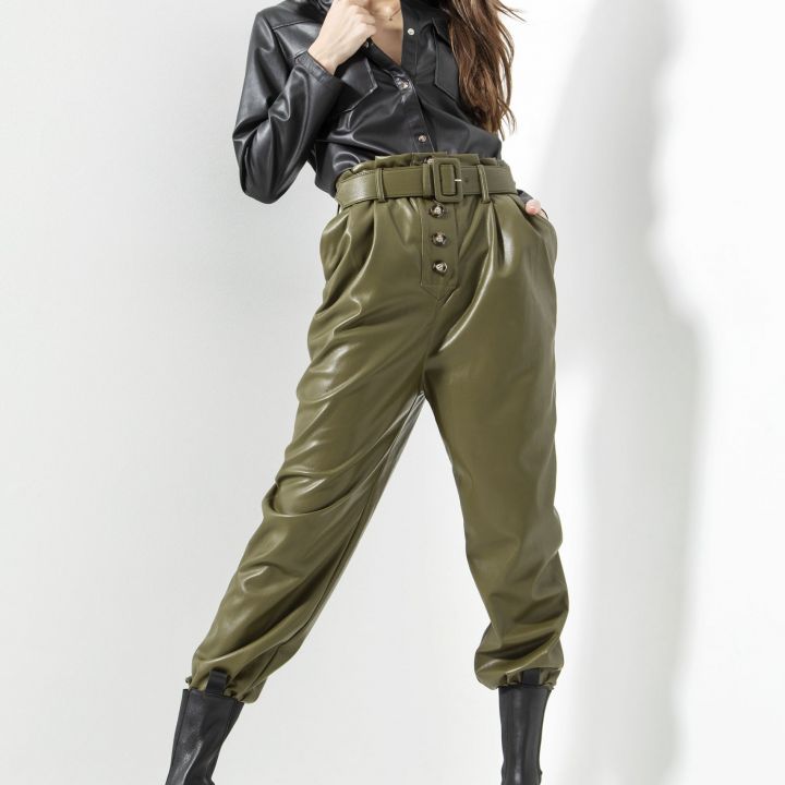 Eine Person posiert selbstbewusst in einem ausgefallenen Modeensemble und trägt ein schwarzes Lederhemd, hoch taillierte olivgrüne Hosen und schwarze Stiefel. Der Look wird durch eine lange Frisur und einen minimalistischen Hintergrund vervollständigt.