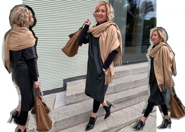 Eine Frau mit lockigem blondem Haar trägt ein schwarzes Kleid, schwarze Leggings und einen hellbraunen Schal. Sie trägt eine braune Handtasche und geht lächelnd und nach vorne blickend die Stufen vor einer gerippten Gebäudefassade hinauf.