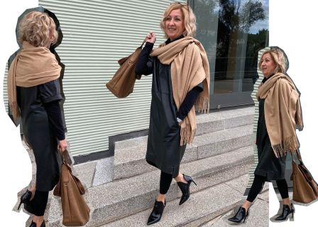 Eine Frau mit lockigem blondem Haar trägt ein schwarzes Kleid, schwarze Leggings und einen hellbraunen Schal. Sie trägt eine braune Handtasche und geht lächelnd und nach vorne blickend die Stufen vor einer gerippten Gebäudefassade hinauf.