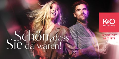 Eine Frau in einer braunen Jacke und ein Mann im grauen Anzug stehen lächelnd zusammen. Der Hintergrund weist einen verschwommenen, farbenfrohen Lichteffekt auf. Text lautet: „Schön, dass Sie da waren!“ und „Kastner & Öhler Inspiriert seit 1873“.