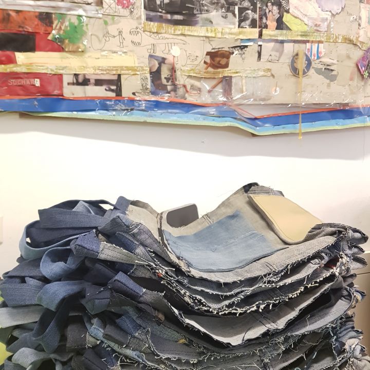 Auf einer grünen Fläche liegt ein Stapel handgefertigter Jeanstaschen. Darüber hängt an der Wand eine farbenfrohe, eklektische Collage aus verschiedenen Materialien, Bildern und Texturen.
