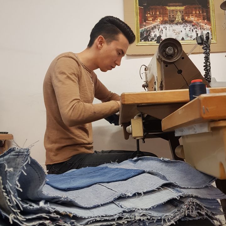 Eine Person näht in einer Werkstatt an einer Maschine, umgeben von gefaltetem Jeansstoff. Im Hintergrund hängen eine Uhr und ein gerahmtes Bild an der Wand.