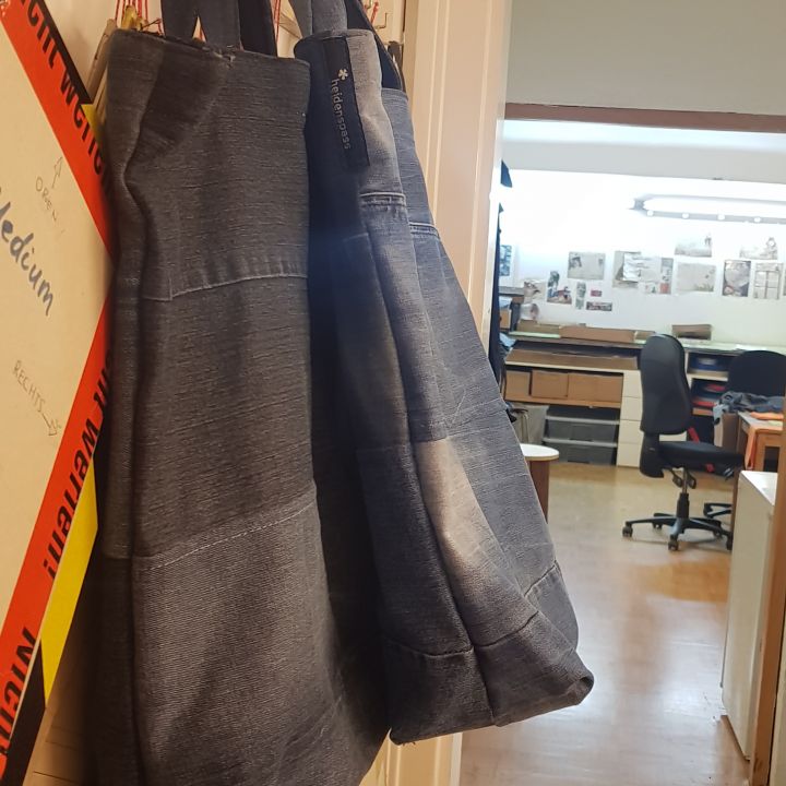 Zwei Jeanstaschen hängen nebeneinander an einer Wand in einem Flur. Im Hintergrund ist ein Büro mit Stühlen, Schreibtischen und verschiedenen ausgestellten Papieren und Tafeln zu sehen. Die Szene ist gut beleuchtet und hat eine warme Atmosphäre.