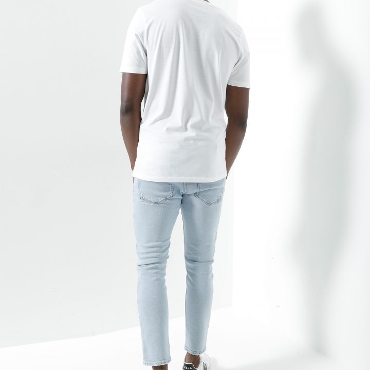Eine Person steht in einem hellen, minimalistischen Raum und trägt ein weißes T-Shirt, hellblaue Jeans und weiße Turnschuhe. Die Person blickt von der Kamera weg und hat eine entspannte Haltung. Der Hintergrund ist eine schlichte weiße Wand.
